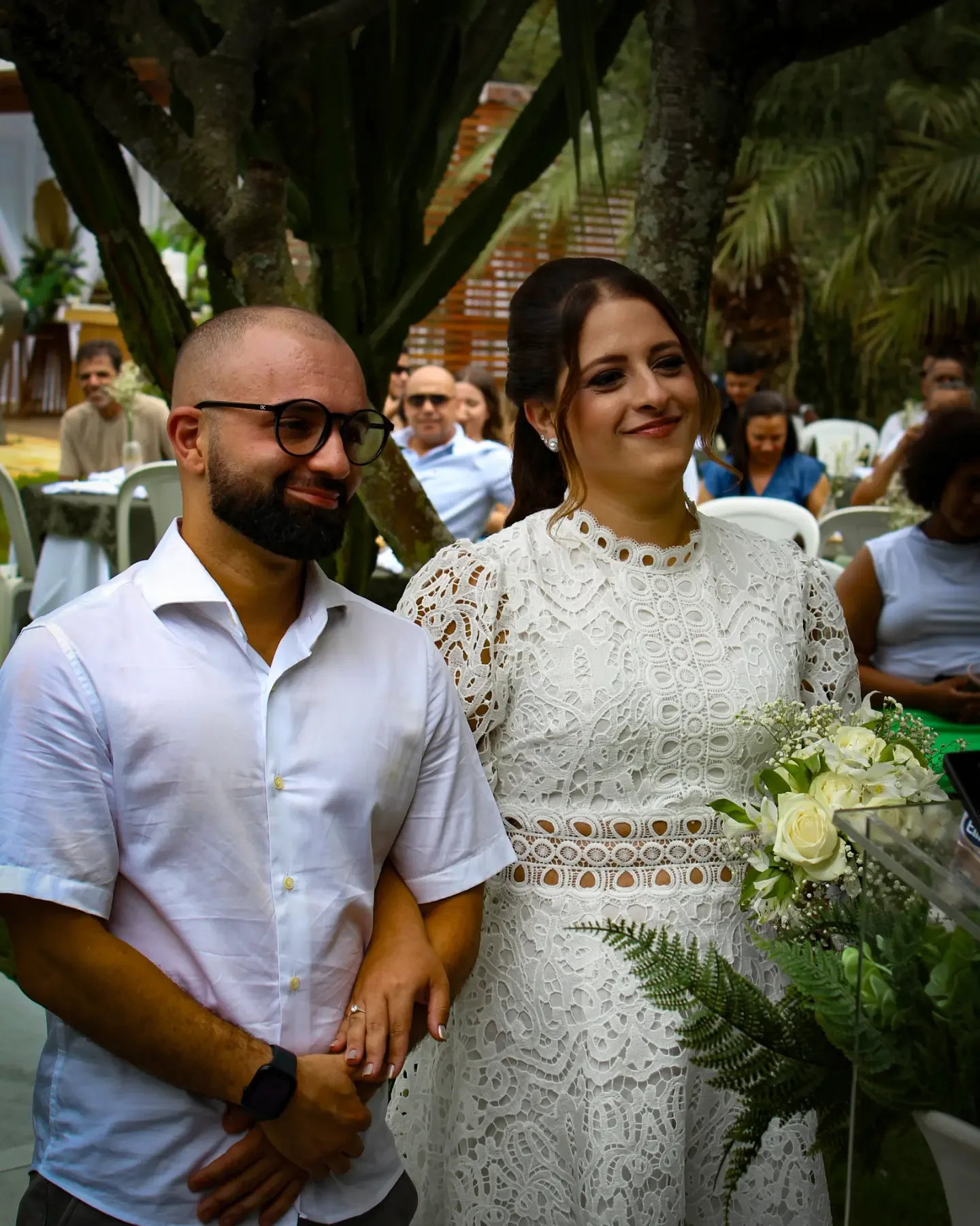 Noivos de braços dados sorriem no jardim; ela segura o buquê.