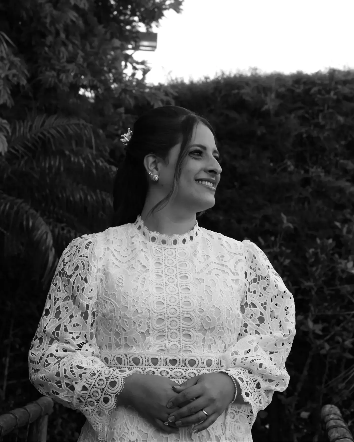 Noiva de vestido de renda sorri e olha para o lado, com as mãos unidas. Foto em preto e branco.