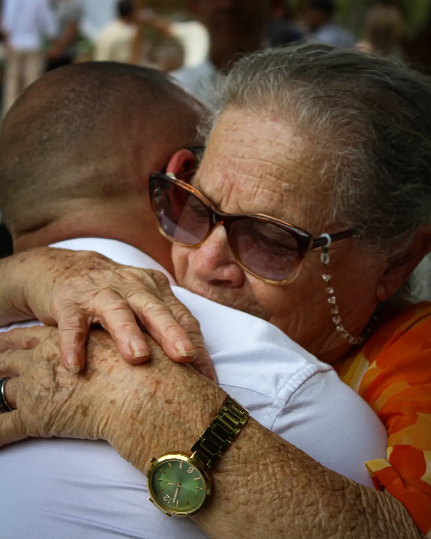 Mulher de óculos escuros abraça o noivo com as duas mãos nas costas dele.