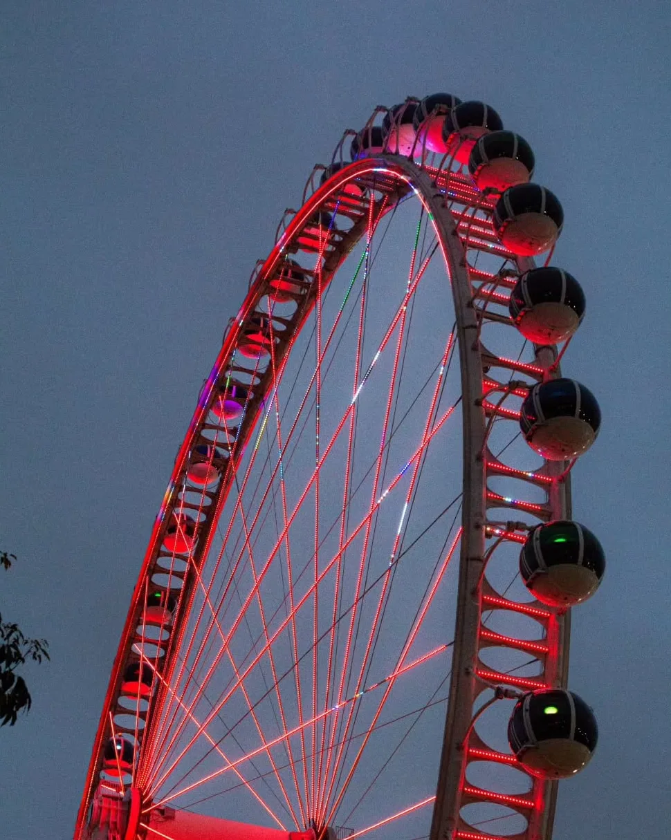 Roda-gigante vista de baixo no fim da tarde, com os raios acesos em vermelho contra o céu azulado.
