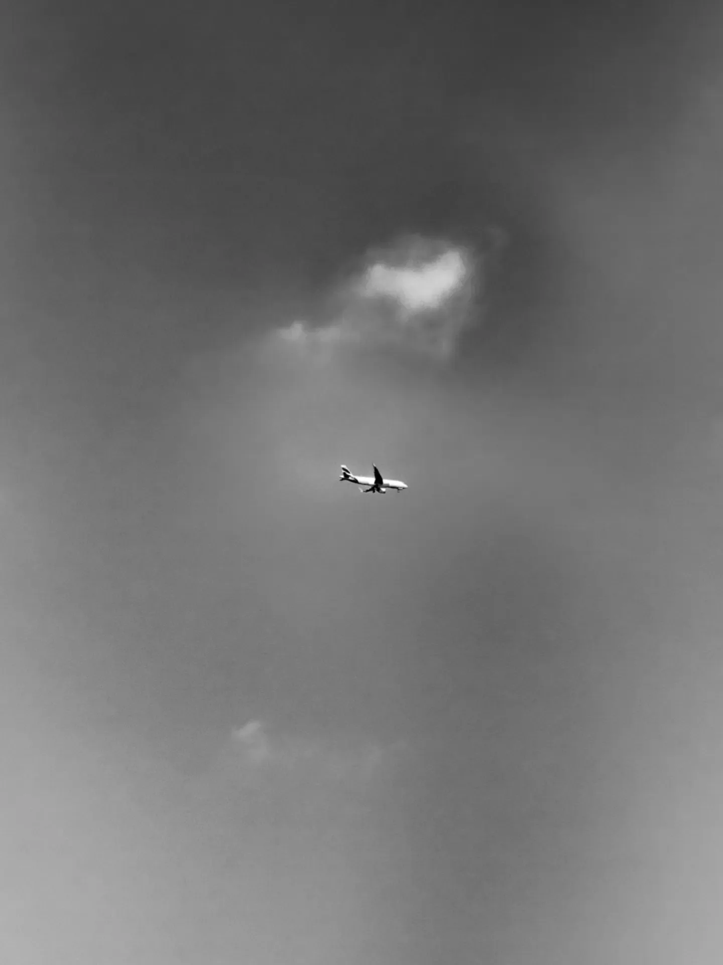 Avião pequeno cruza o céu cinza ao lado de uma nuvem. Foto em preto e branco.