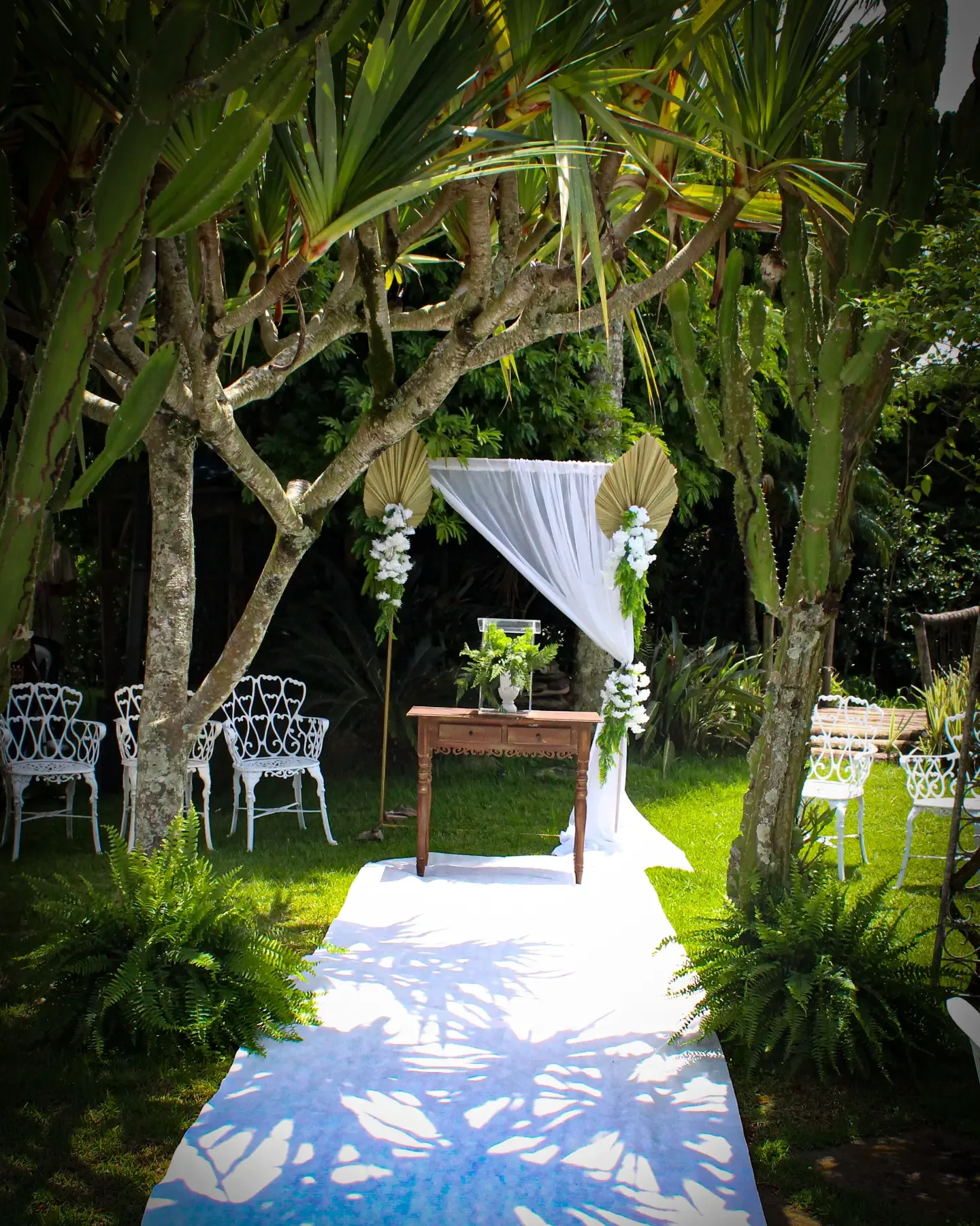 Corredor branco sob as árvores leva a um altar com tecido, mesa e cadeiras no jardim.