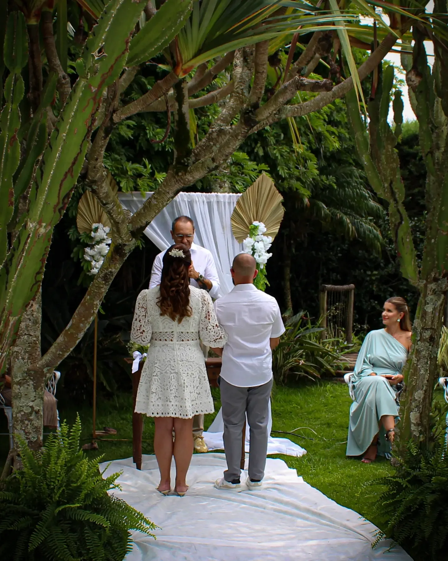 Noivos de costas diante do altar, vistos do fim do corredor, entre as árvores.