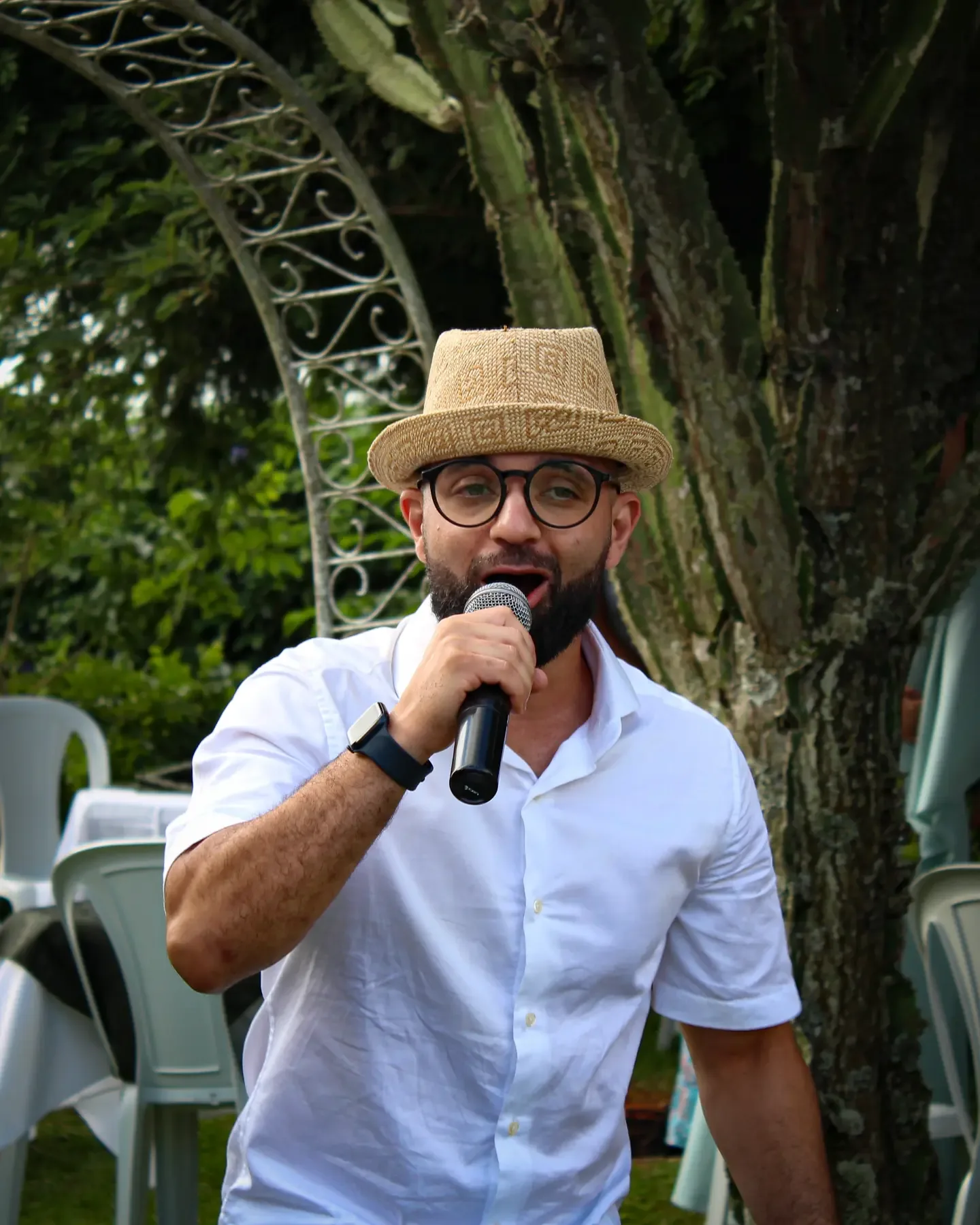 Homem de chapéu de palha e camisa branca fala ao microfone no jardim.