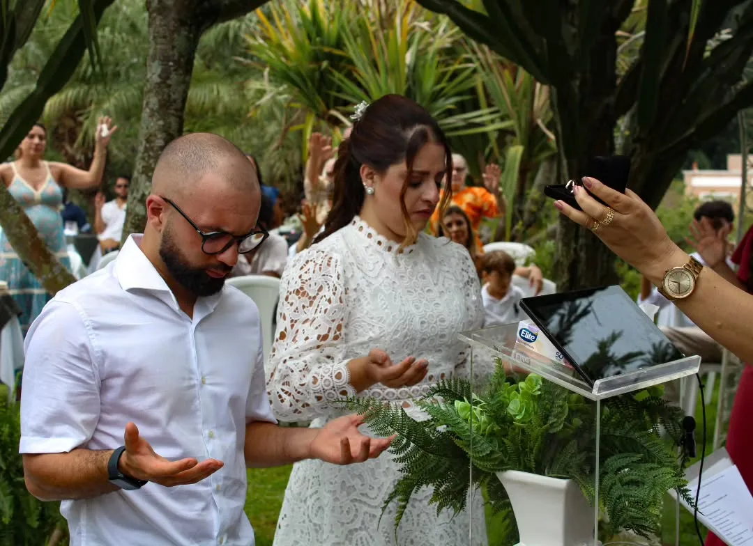 Noivos com as mãos abertas diante de um tablet apoiado em um púlpito, cercados por convidados.
